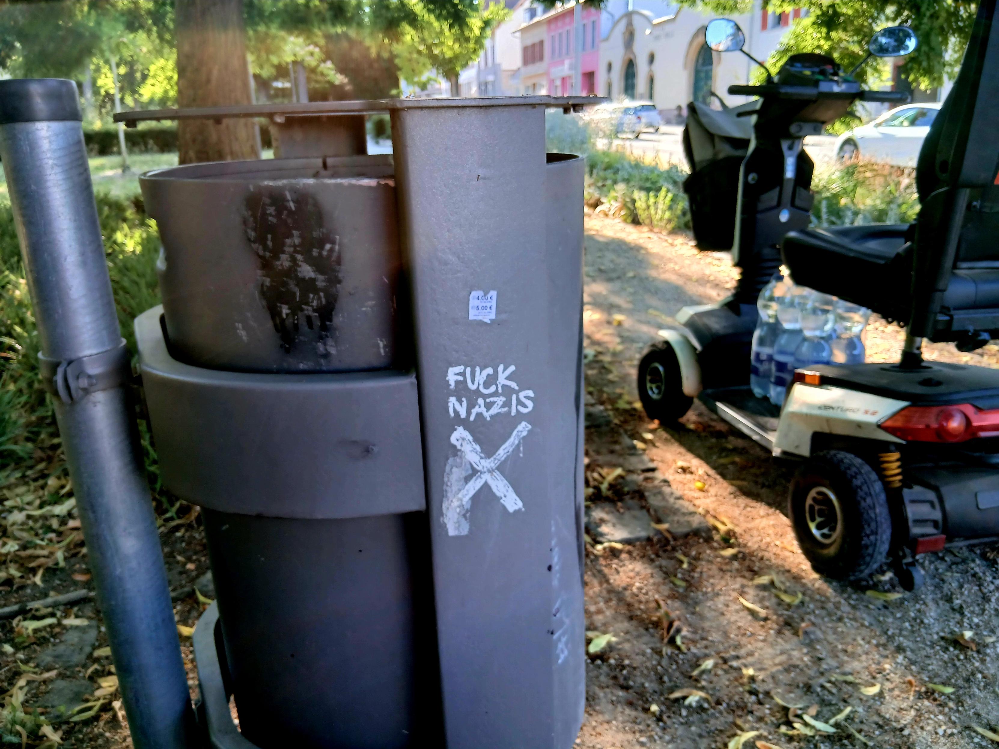 The scoot-chair is parked next to a trash can emblazoned with FUCK NAZIS, and the Shitter logo, which is a sans-serif swastika.