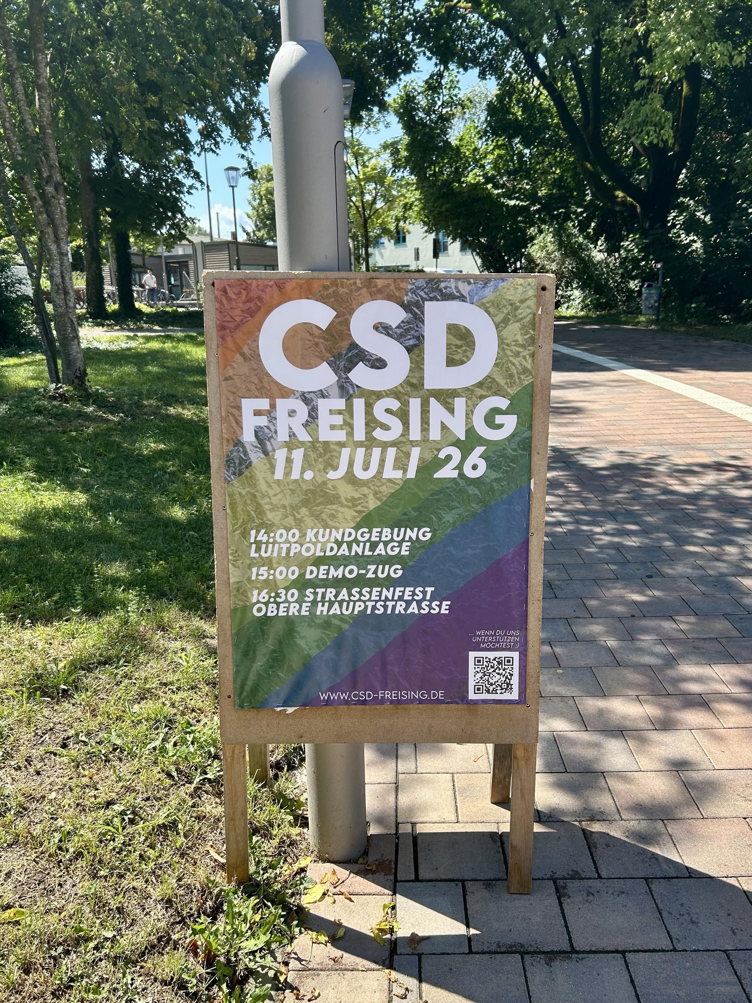 The center of the photo is a wooden sign with a colorful poster announcing the CSD (Christopher Street Day) in Freising on July 11. The poster also lists the schedule: a gathering at 14:00, a parade at 15:00, and a street festival at 16:00. The sign is leaning against a lamppost. On its left side, there is grass, on the right side a walkway. In the upper part of the photo are trees in the background.