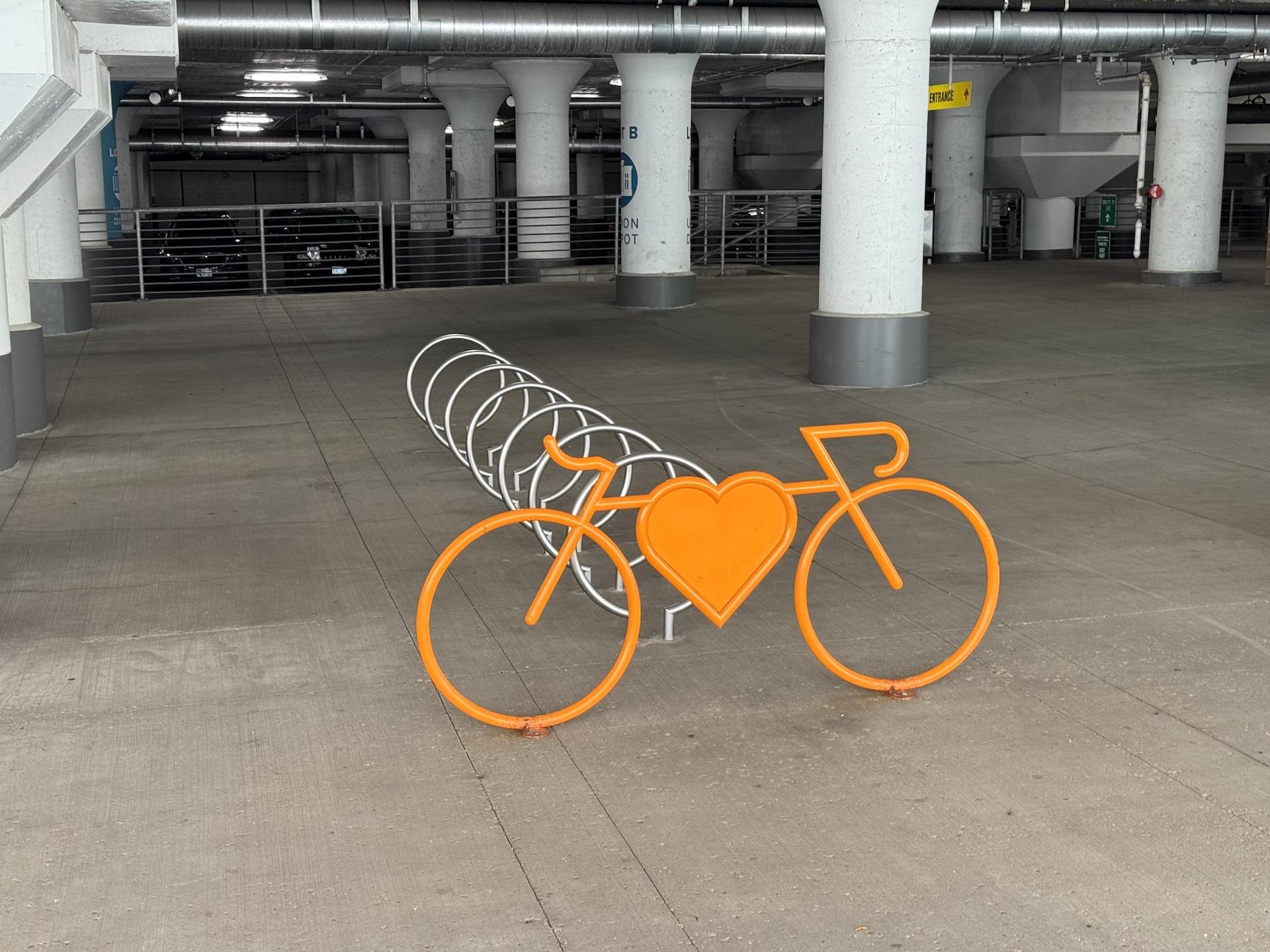Photo of a bicycle rack on the edge of a covered parking area. The bicycle rack is comprised of a stylized bicycle shape with a large heart in the center, all orange, and spaced behind that are seven large silver metal hoops spaced out. The photo perspective shows them fading to the left. The bicycle rack is on a concrete surface and a few rows of white columns are seen behind the rack.
