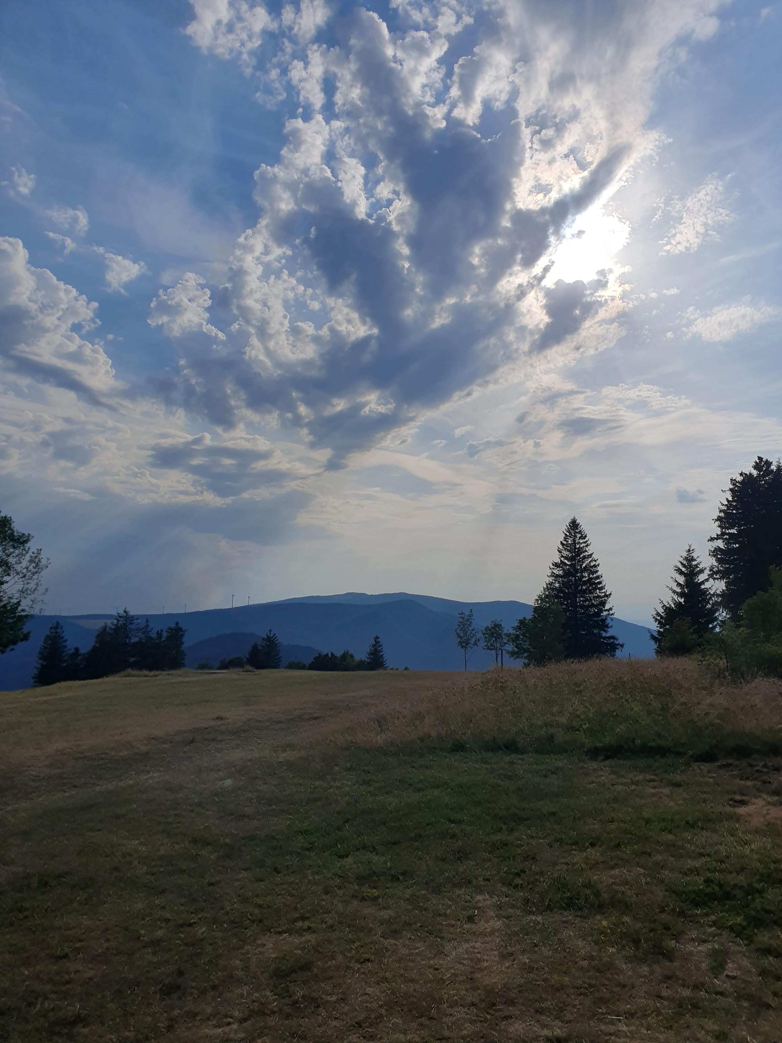 Blick von einer grasbewachsenen Bergkuppe mit vereinzelten Bäumen hinüber zu weiteren bewaldeten Bergen. Man kann im Dunst ein paar Windräder entdecken. Die Sonne scheint, wird aber gerade dramatisch von einer Eolke bedeckt.