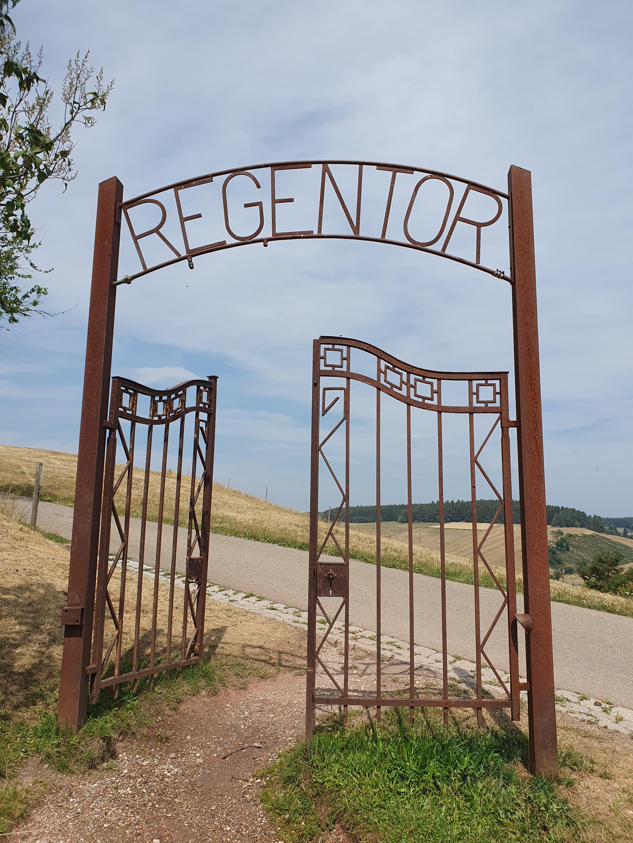 Neben einer schmalen Straße steht ein hohes Tor - nur das Tor - aus verrostetem Metall. Über den Türen, von denen eine geöffnet ist, steht "Regentor". Im Hintergrund sieht man hügelige Landschaft mit trockenen Weiden und dunklen Wäldern.