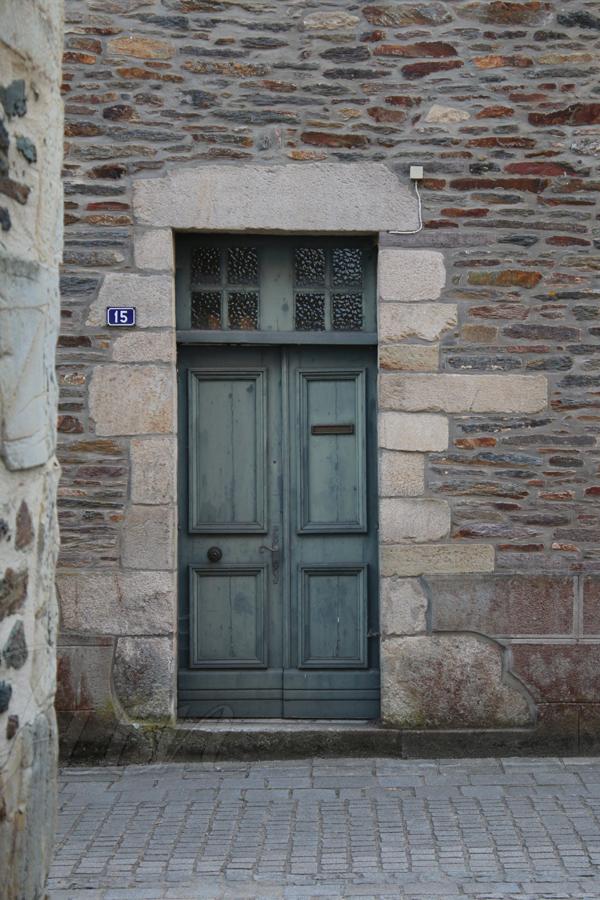 A faded pine green wooden door is framed in cream granite stones in a wall of grey and ocre slate stones. The double door has four rectangular moldings and a high slot for slipping letters through. The left side of the door has a flat circular door knob and an old fashioned sculpted door handle. In the framing, there are also two square mullioned windows above the door. A paved alley is passing by, in front of the door.