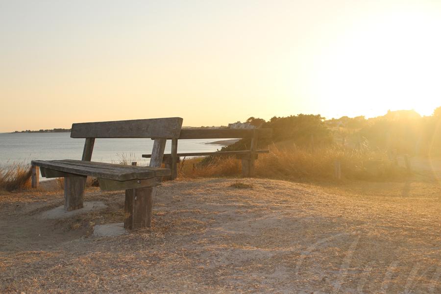 Two wooden benches face the ocean on the top of a sand dune. There are some dry shrubs around them. The photograph is taken against the light while the Sun is very low, near sunset. 
––––––
Shot with Canon 70D + EFS 18‑55mm f/3.5‑5.6 IS2 on August 2022.
29mm – 1/250s – f/7,1 – ISO 100