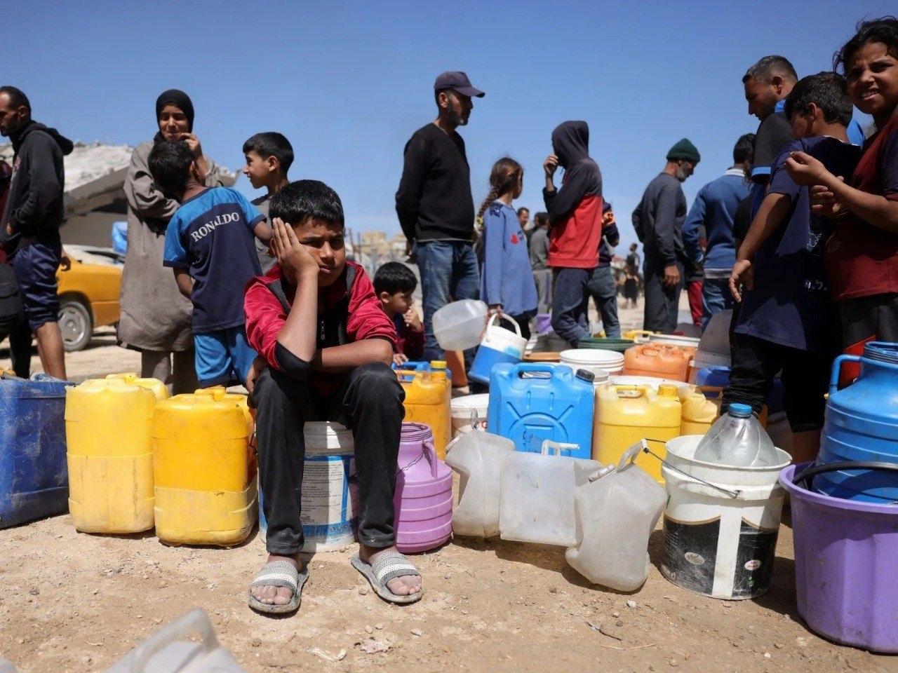 A line of children waits for the water truck under the scorching sun, their containers empty.