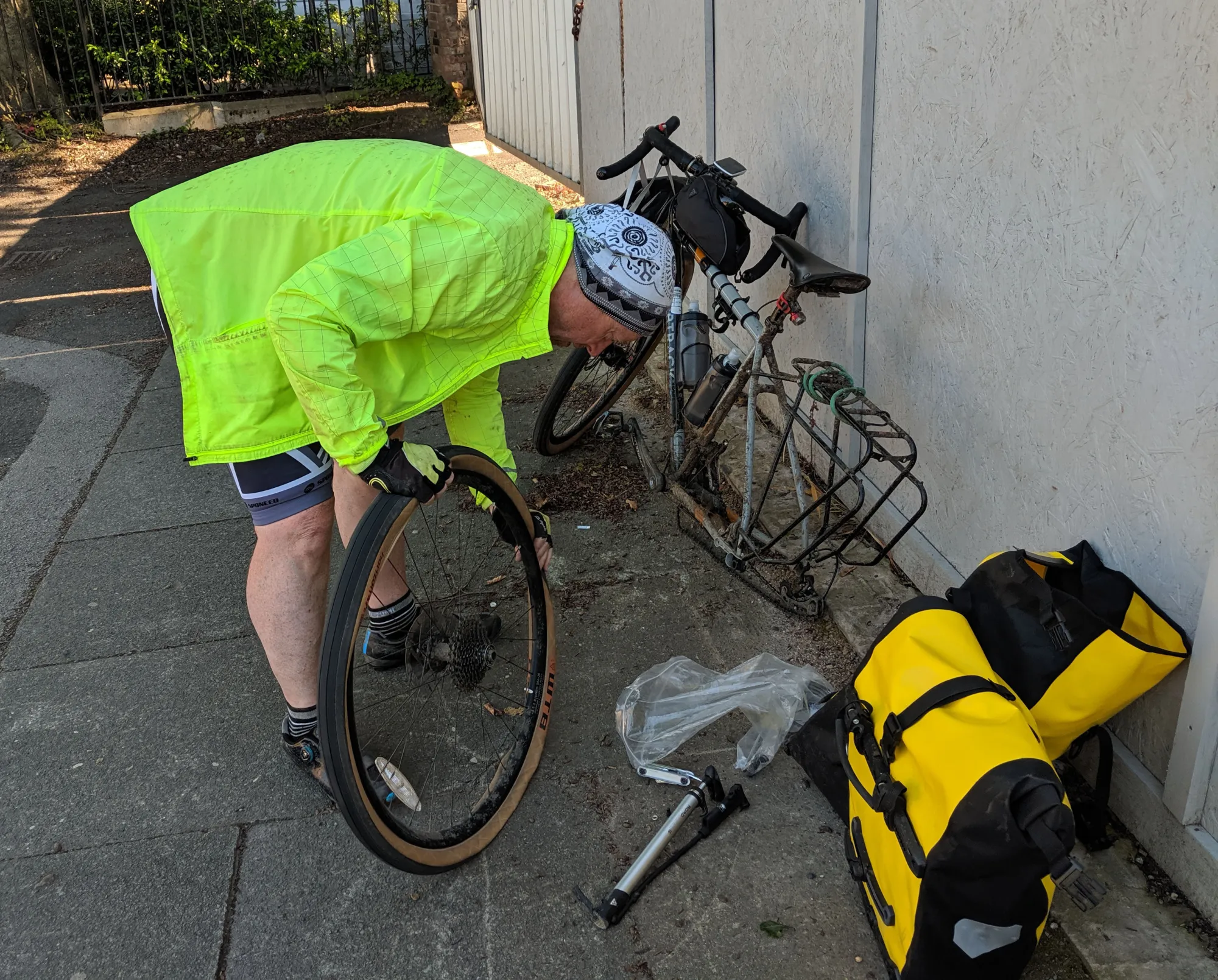 Cyclist in yellow windbreaker fixing a flat tire. The bicycle is behind him, resting upright with the rear wheel removed. In the foreground right, two bright yellow pannier bags are resting on the pavement.
