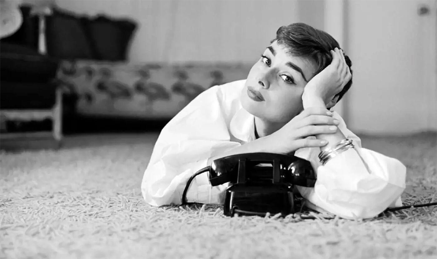 Audrey lies on her belly, wearing a white men's shirt and some bangles, holding her head in her hand, as she waits for the corded black telephone in front her to ring.