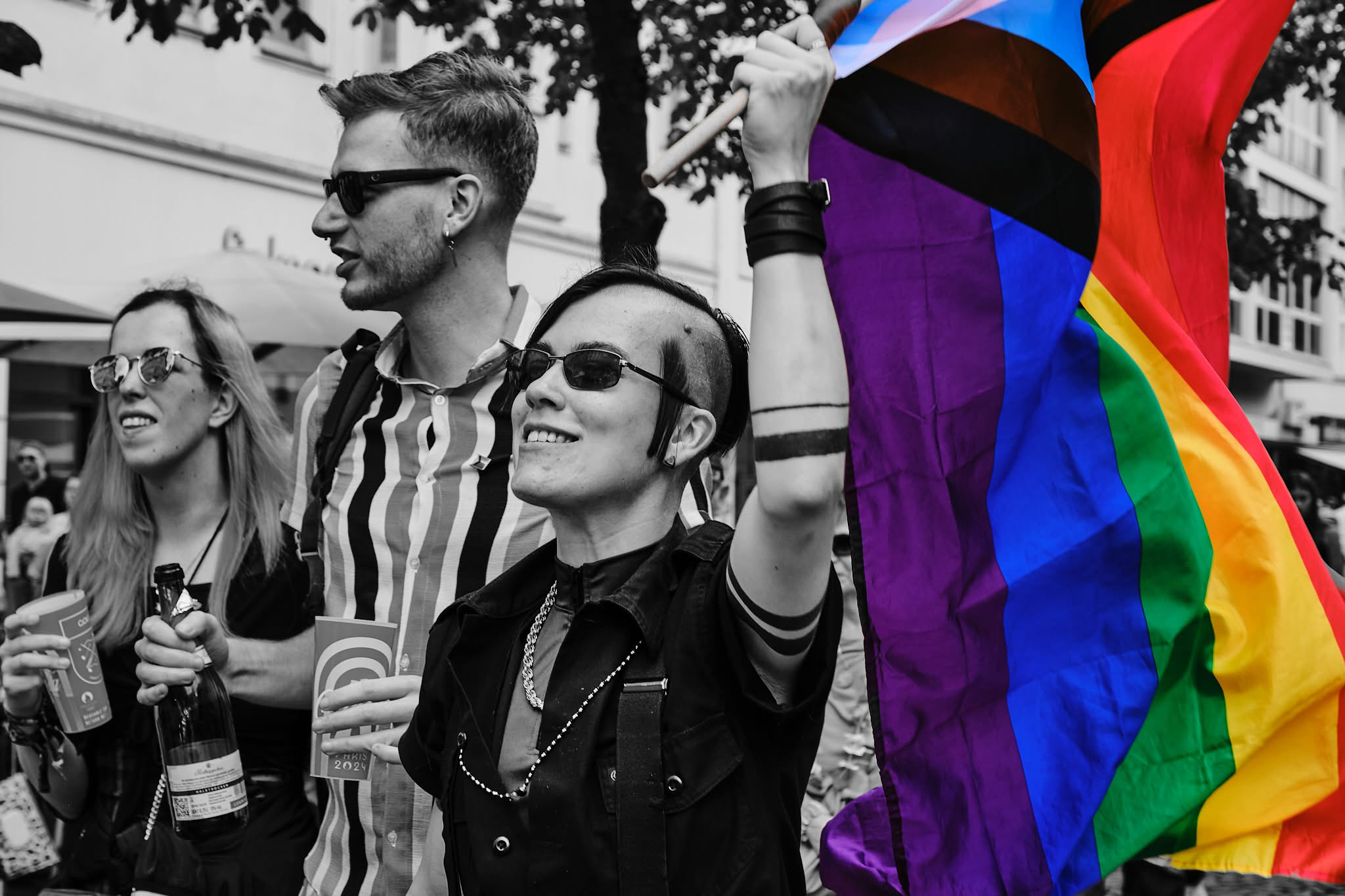 A high-contrast photo rendered mostly in black and white, featuring a selectively colored Progress Pride flag.
In the foreground, a person with dark, short hair, sideburns, and small dark sunglasses smiles while holding up a flagpole. The Progress Pride flag unfurls to the right, vibrant with saturated rainbow stripes alongside the light blue, pink, white, brown, and black chevron pattern. The person holding the flag wears a black short-sleeved button-up shirt, a silver chain necklace, black band tattoos around their forearm, and a wide leather wristband.
Beside them, a man in dark sunglasses and a vertical-striped shirt looks to the side. To the far left, a person with long light-colored hair and sunglasses holds a glass and a bottle while smiling. The background shows an outdoor street setting with trees and buildings.